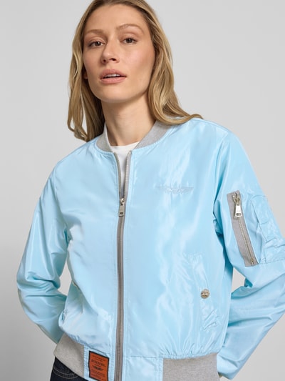 Frauenmodel trägt Bomberjacke mit Label-Patch und Reißverschlusstasche am Ärmel von Original Bombers in Hellblau mit grauen elastischen Abschlüssen, Lockerer-Schnitt, Stehkragen, silberfarbener Reißverschluss, Logo-Stitching, seitliche Eingrifftaschen mit Druckknopfverschluss, Detailaufnahme Oberkörper.
