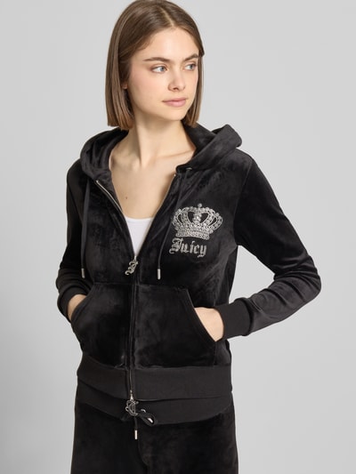 Kobiece model prezentuje Sweatjacke mit Ziersteinbesatz Modell 'GOTHIC CROWN ROBERTSON' od Juicy Couture, czarny materiał, elastyczny, krój slim fit, ozdobne kamienie, kaptur z troczkiem, dwukierunkowy zamek, zbliżenie na górną część ciała, ujęcie z przodu.