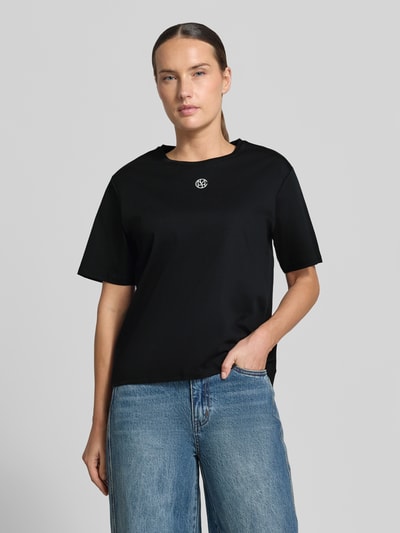 Kobiece modelka nosi czarny Damski t-shirt Regular Fit Model 'Melea' od MSCH Copenhagen z bawełny, nadruk z logo, okrągły dekolt, Długość tyłu w rozmiarze XS/S: 57 cm, zdjęcie całej sylwetki z przodu.