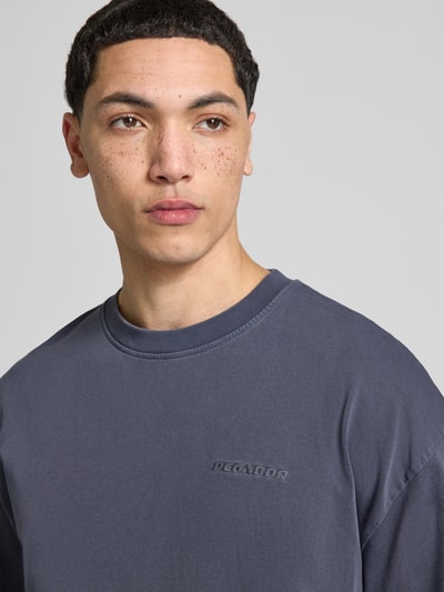 Męski model trägt Oversized T-Shirt mit Logo-Stitching von Pegador aus bawełna, krój oversized fit, okrągły dekolt, długość tyłu w rozmiarze M: 73 cm, wyhaftowane logo, kolor szary, gładka faktura, zbliżenie na górną część ciała.