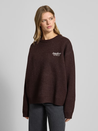 Frauenmodel trägt Oversized Strickpullover mit Rückenmotiv Modell 'Wine & Vibe' von ANOTHER COTTON LAB, Rundhalsausschnitt, Label-Stitching, lockere Passform in dunklem Braun, weiche Textur, Damen Strickpullover, Ganzkörperansicht von vorne.
