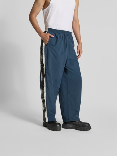 Männermodel trägt Trackpants mit elastischem Bund von VETEMENTS in Blau mit Galonstreifen, Eingrifftaschen und Mesh-Futter, Ganzkörperansicht von vorne. Innenbeinlänge 69 cm, Bundweite 44 cm bei Größe M, Regular-Fit.