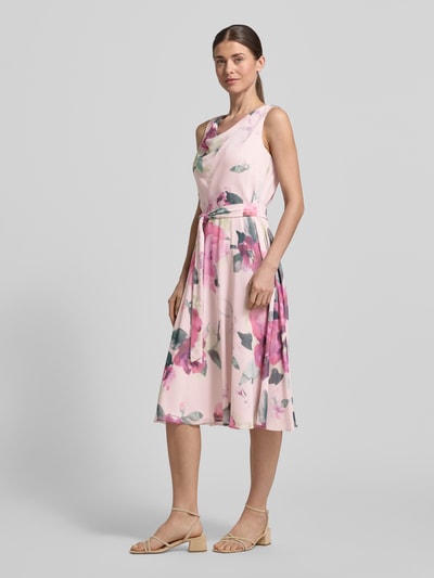 Frauenmodel trägt Knielanges Cocktailkleid mit Bindegürtel von Vera Mont aus Chiffon, Taillierter Schnitt, Wasserfall-Ausschnitt, florales Muster, Ganzkörperansicht von vorne. Rückenlänge bei Größe 36: 104 cm, Brustweite bei Größe 36: 40 cm.