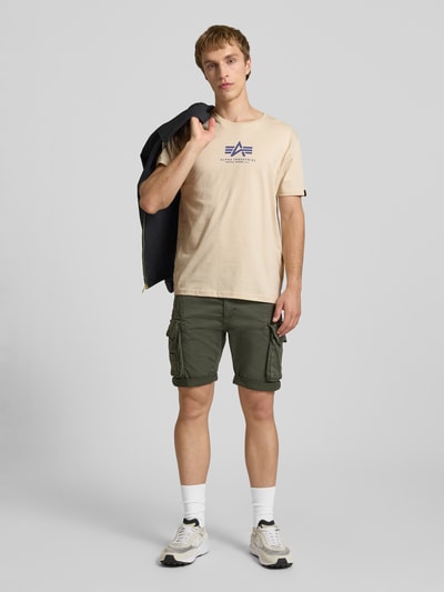 Männermodel trägt T-Shirt mit Logo und Rundhalsausschnitt von Alpha Industries in Beige, Reine Baumwolle, Regular-Fit, Rückenlänge 67 cm, Ganzkörperansicht von vorne.