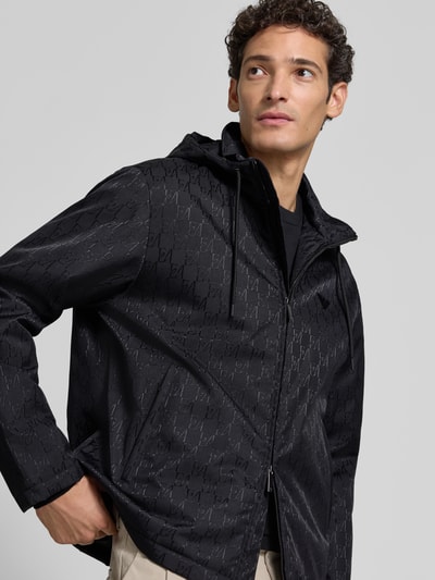 Männermodel trägt Regular Fit Jacke mit Allover-Logo-Print von Emporio Armani in Schwarz mit Kapuze, Zweiwege-Reißverschluss, Nahaufnahme Oberkörper. Kontext: Damen Jacke, Logo-Applikation, Tunnelzug, Eingrifftaschen, Rückenlänge 61 cm, Schulterbreite 47 cm, Brustweite 58 cm, Ärmellänge 65 cm.