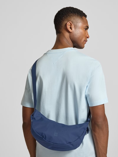 Männermodel trägt Umhängetasche mit Label-Detail von Gotbag in Blau mit verstellbarem Schulterriemen, Ganzkörperansicht Rückansicht. Breite 32 cm, Länge 19 cm, Tiefe 10 cm, Länge Schulterriemen 82 cm, Hauptfach mit Reißverschluss, Innenfach mit Reißverschluss.