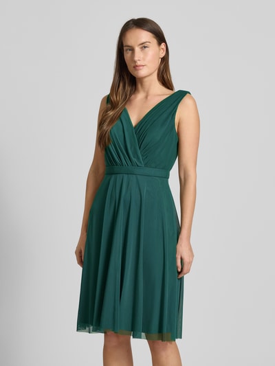 Frauenmodel trägt Cocktailkleid mit V-Ausschnitt von TROYDEN COLLECTION in Dunkelgrün mit Raffungen, Taillengürtel und blickdichtem Unterkleid, Ganzkörperansicht von vorne. Taillierter Schnitt, Nahtreißverschluss, Rückenlänge 79 cm, Brustweite 24 cm.