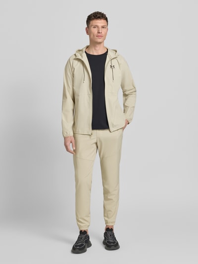 Męski model nosi Regular Fit Jacke mit Logo-Print Modell 'Vibe' od Under Armour, beżowy kolor, długa bluza z kapturem, krój regular fit, materiał elastyczny, kieszenie, zamek, widok całej sylwetki z przodu.