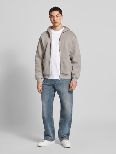 Męski model nosi Sweatjacke mit Känguru-Tasche od REVIEW, kolor szary, relaxed fit, bawełniany miks, widoczny zamek, dżinsy, białe buty, długość tyłu 68 cm, długość rękawa 62 cm, widok całej sylwetki z przodu.