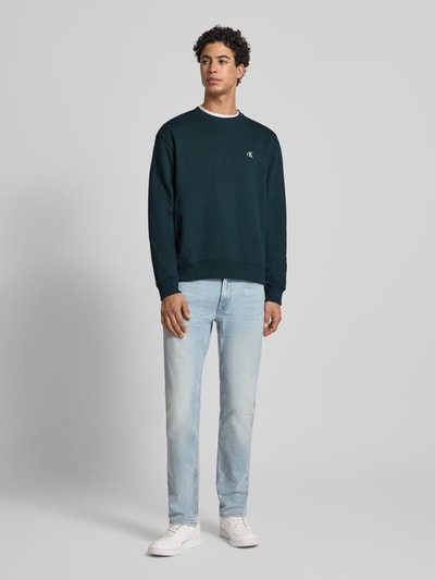 Männermodel trägt Sweatshirt aus reiner Baumwolle von Calvin Klein Jeans in Dunkelgrün mit Label-Stitching und gerippten Abschlüssen, Ganzkörperansicht von vorne.