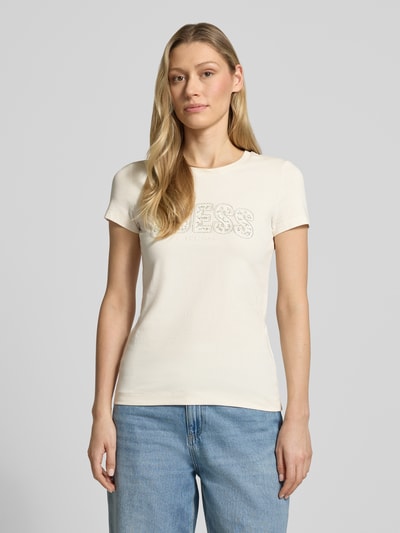 Frauenmodel trägt Slim Fit T-Shirt mit Strasssteinbesatz von Guess aus elastischem Baumwoll-Mix, Logo-Detail, Rundhalsausschnitt, Farbe Creme, Rückenlänge 59 cm bei Größe S, Ganzkörperansicht von vorne.