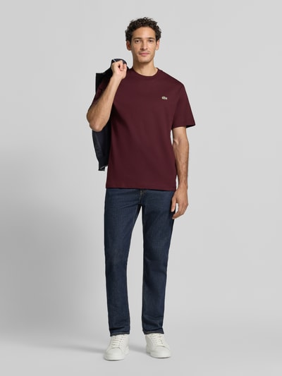 Männermodel trägt Regular Fit T-Shirt aus reiner Baumwolle von Lacoste in Dunkelrot, kombiniert mit Jeans, Ganzkörperansicht von vorne.