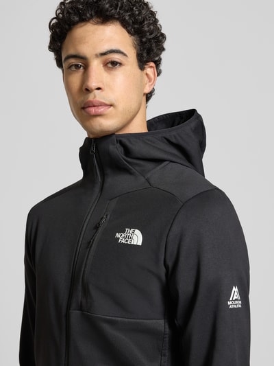 Männermodel trägt Sweatjacke mit Napoleontasche Modell 'MOUNTAIN ATHLETICS' von The North Face, Schwarz, Label-Details, Nahaufnahme Oberkörper.