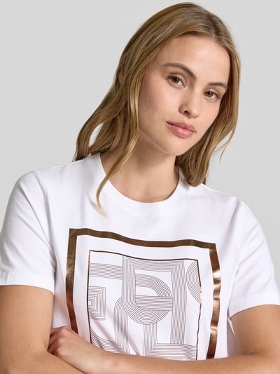 Frauenmodel trägt T-Shirt mit Motiv-Print von comma in Weiß mit geometrischem, glänzend-bronze-farbenem Muster, Rundhalsausschnitt, Regular Fit, Baumwoll-Mix mit Modal-Anteil, Nahaufnahme Oberkörper.