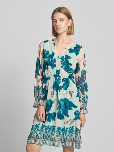 Frauenmodel trägt knielanges Kleid im Allover-Look Modell 'Ja44milaa' von ZABAIONE, Chiffon, A-Linie, V-Ausschnitt, Wickel-Optik, florales Muster in Blau- und Beigetönen, zarte Struktur, Ganzkörperansicht von vorne.