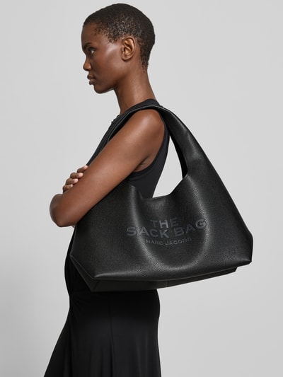 Frauenmodel trägt Shoulder Bag aus echtem Rindsleder von Marc Jacobs in Schwarz mit Logo-Detail, Hauptfach mit Magnetverschluss, herausnehmbare Innentasche mit Reißverschluss, Tragehenkel, Maße ca. 42 cm Breite, 23 cm Länge, 20 cm Tiefe, Ganzkörperansicht von der Seite.