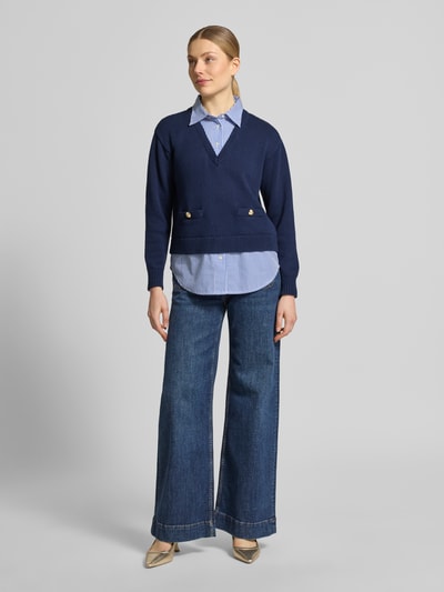 Modelka kobieca nosi Regular Fit Strickpullover aus reiner Baumwolle von Lauren Ralph Lauren w granatowym kolorze, tekstura dzianiny, paskowany element, logo, długi rękaw, dekolt V, dół i rękawy zakończone ściągaczem, widoczna koszula pod spodem, widok całej sylwetki z przodu.