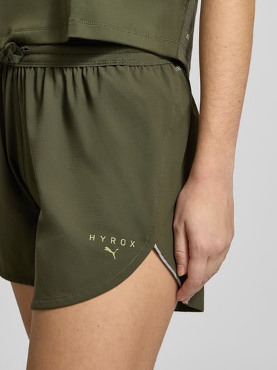Frauenmodel trägt PUMA x HYROX - Shorts mit Label-Print in Olivgrün, lockerer Schnitt, elastischer Bund mit Tunnelzug, Nahaufnahme Oberschenkel.