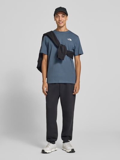 Männermodel trägt T-Shirt mit Label-Print Modell 'Evolution' von The North Face in Blau, Baumwoll-Mix, dunkle Jogginghose, Ganzkörperansicht von vorne, Herren T-Shirt Regular Fit, Rückenlänge bei Größe M 73 cm.