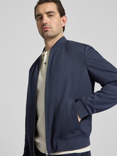 Męski model nosi Jacke mit Reißverschluss od MCNEAL, niebieski materiał z domieszką wiskozy i elastycznym wykończeniem, krój regular fit, ściągacze, stójka, kieszenie boczne, widoczne detale konstrukcji, szerokość w rozmiarze M: 57 cm, długość tyłu: 63 cm, szerokość ramion: 46 cm, długość rękawa: 67 cm, całkowity widok górnej części ciała z przodu.