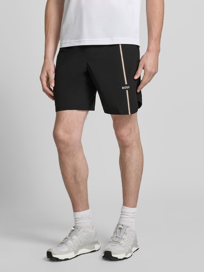 Mężczyzna nosi czarne Regular Fit Sportshorts mit Logo-Details model 'SH_TOC GAME 8 INCH' von BOSS Green, widoczne białe i beżowe akcenty, Ganzkörperansicht von vorne.