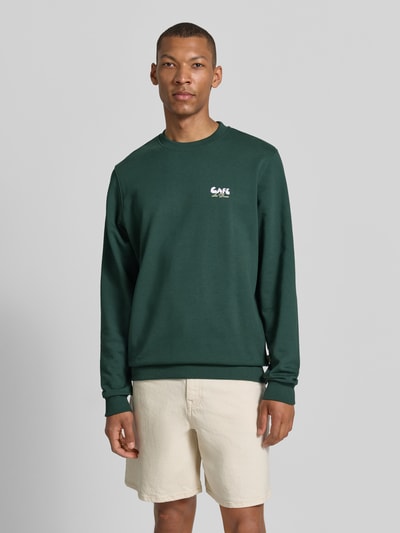Männermodel trägt Sweatshirt mit Label-Details und Rundhalsausschnitt von Les Deux, dunkelgrün, Baumwoll-Elasthan-Mix, Regular-Fit, Ganzkörperansicht von vorne, Rückenlänge 66 cm, Ärmellänge 70 cm, Größe M.