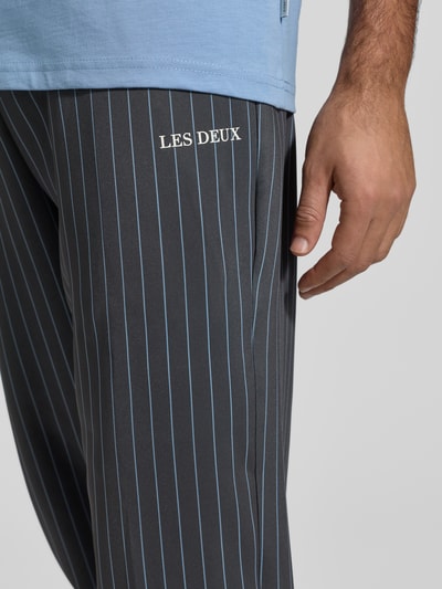 Mężczyzna nosi Stoffhose mit elastischem Bund Modell 'Ballier' von Les Deux, ciemne spodnie w prążki, materiał z domieszką stretchu, z widocznym nadrukiem marki, kieszenie francuskie, elastyczny pas z tunelem, luźny krój, zbliżenie na udo z przodu.