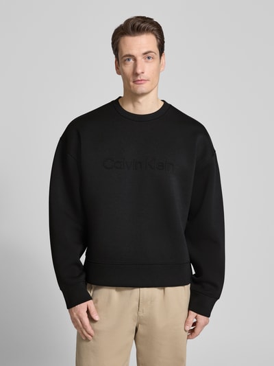 Męski model nosi Bluza Sweatshirt mit Rundhalsausschnitt und Logo od CK Calvin Klein, czarny kolor, mieszanka materiałów z dodatkiem wiskozy, krój regular fit, okrągły dekolt, logo, długość tyłu w rozmiarze M: 61 cm, długość rękawa w rozmiarze M: 59 cm, widok całej sylwetki z przodu.