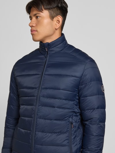 Męski model nosi kurtkę Regular Fit Jacke in Stepp-Optik Model 'Kingsley' od JACK & JONES, granatowy kolor, stójka, zamek błyskawiczny, wpuszczane kieszenie, długość tyłu w rozmiarze M 72 cm, szerokość ramion w rozmiarze M 48 cm, szerokość klatki piersiowej w rozmiarze M 51 cm, długość rękawów w rozmiarze M 68 cm, z efektem pikowania, zdjęcie w półzbliżeniu od przodu.