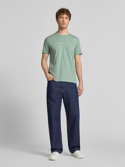 Männermodel trägt T-Shirt mit Label-Detail von Alpha Industries in Grün, Reine Baumwolle, Gerader-Schnitt, Rundhalsausschnitt, Ganzkörperansicht von vorne.