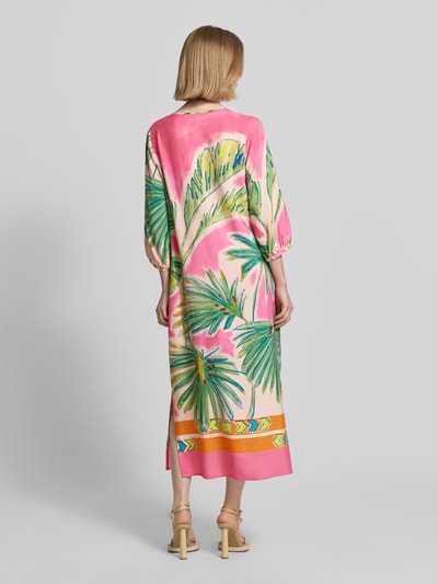 Frauenmodel trägt Tunikakleid aus Viskose mit Tunikakragen und Quasten von Emily Van den Bergh in Rosa, Grün und Gelb, Loose Fit, Midilänge, 3/4-Arm, Seitenschlitze, fließende Textur, Ganzkörperansicht Rückansicht. Rückenlänge 124 cm, Brustweite 46 cm, Ärmellänge 43 cm.