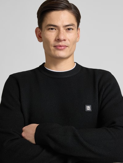 Mężczyzna nosi Regular Fit Strickpullover mit Schurwoll-Anteil Modell 'SETAL' od HUGO, czarny, dzianina, naszywka z logo, zbliżenie na górną część ciała.