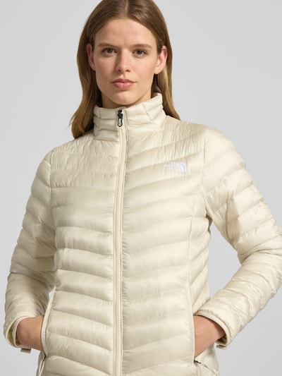Kobiece model nosi kurtka pikowana z stójką i wyhaftowanym logo Steppjacke mit Stehkragen und Label-Stitching von The North Face, krój regular fit, zamek błyskawiczny, kieszenie zapinane na zamek błyskawiczny, długość tyłu w rozmiarze S: 62 cm, szerokość ramion w rozmiarze S: 38 cm, szerokość klatki piersiowej w rozmiarze S: 46 cm, długość rękawów w rozmiarze S: 64 cm, jasny kolor, gładka tekstura, z przodu, nahaufnahme oberkörper.