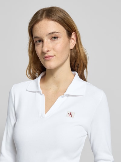 Kobiece model prezentuje Longsleeve mit Label-Detail od Calvin Klein Jeans, biały, krój slim fit, bawełna z elastanem, kołnierzyk polo, dekolt V, detal z logo, długość tyłu w rozmiarze S: 57 cm, zbliżenie na górną część ciała.