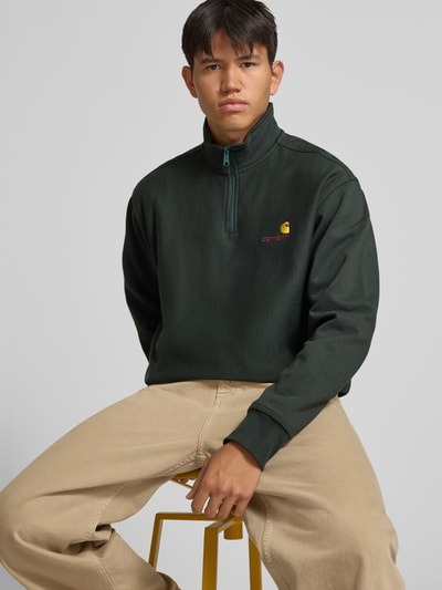 Männermodel trägt Relaxed Fit Sweatshirt aus elastischem Material-Mix von Carhartt Work In Progress, dunkles Grün mit Label-Detail, Troyer-Kragen, elastische Abschlüsse, Ganzkörperansicht von vorne.