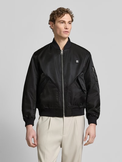 Männermodel trägt Regular Fit Bomberjacke mit Label-Applikation Modell 'Badam2611' von HUGO, schwarz, Reißverschluss, College-Kragen, Ganzkörperansicht von vorne.