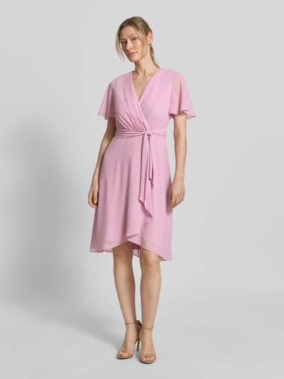 Vrouwenmodel draagt Knielanges Cocktailkleid in Wickel-Optik van Christian Berg Cocktail in lichtroze chiffon, a-lijn, v-hals, knielengte, bindriem, ruglengte maat 36: 100 cm, borstwijdte: 33 cm, mouwlengte: 22 cm. Volledige lichaamsaanzicht van voren.