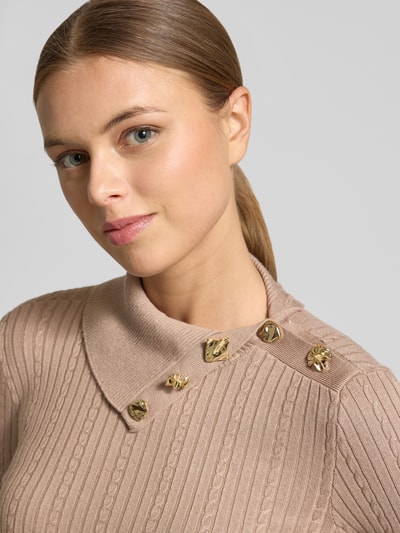 Modelka nosi Slim Fit Longsleeve aus Viskose-Mix Modell 'Sicilla' od Guess, krój slim fit, optyka dzianinowa, stójka, złote ozdoby, kolor beżowy. Zbliżenie na górną część ciała.