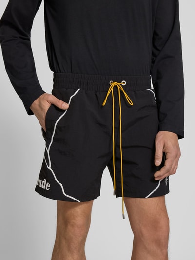 Männermodel trägt schwarze Shorts mit Label-Stitching von RHUDE, gelber Tunnelzug, weiße Naht, seitliche Eingrifftaschen, Nahaufnahme Oberkörper.