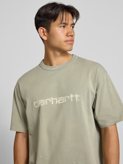 Męski model nosi Regular Fit T-Shirt aus reiner Baumwolle Modell 'Painter' von Carhartt Work In Progress w kolorze jasnoszarym z detalem z logo, kontekst: Męski t-shirt Carhartt Work In Progress, bawełna, krój regular fit, Model 'Painter', detal z logo, Długość tyłu w rozmiarze M: 72 cm, nahaufnahme górnej części ciała.