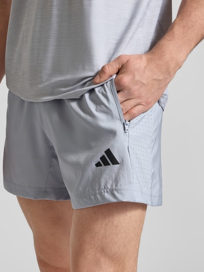 Männermodel trägt Shorts mit Label-Print von adidas Training in Hellgrau, Lockerer-Schnitt, Reißverschlusstaschen, Nahaufnahme Oberschenkel.