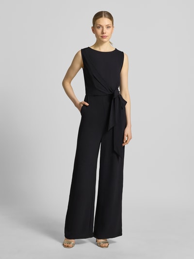Frauenmodel trägt Jumpsuit mit Bindegürtel Modell 'Jessica' von GUIDO MARIA KRETSCHMER WOMAN in Schwarz, Taillierter Schnitt, U-Boot-Ausschnitt, Wickel-Optik, rückseitiger Reißverschluss, glatte Textur, Ganzkörperansicht von vorne. Rückenlänge 36 cm, Brustweite 38 cm, Gesamtlänge 148 cm, Innenbeinlänge 77 cm bei Größe 36.