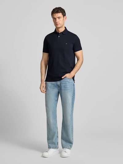Männermodel draagt Regular Fit Poloshirt met Logo-Stitching van Tommy Hilfiger, mix van katoen en elastaan, polokraag, knoopsluiting, heren, ruglengte maat M 73 cm, donkerblauw, lichte jeans, witte sneakers, neutrale achtergrond. Ganzkörperansicht van voren.
