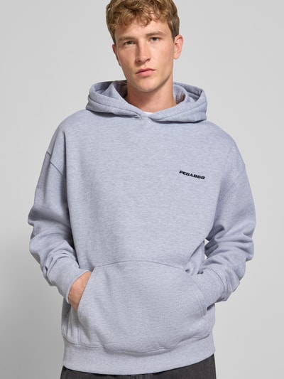 Männermodel trägt Oversized Hoodie aus Baumwoll-Mix von PEGADOR in Hellgrau mit Logo-Stickerei, Kängurutasche, Ganzkörperansicht von vorne.