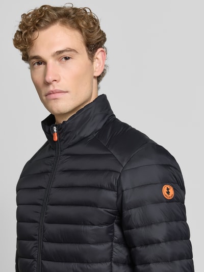 Männermodel trägt gewatteerd jack Model 'ALEXANDER' van Save The Duck in zwart met horizontale doorgestikte naden, slim fit, opstaande kraag, tweewegritssluiting, ritszakken, ruglengte maat M 67 cm, schouderbreedte 47 cm, borstwijdte 57 cm, mouwlengte 68 cm, Nahaufnahme bovenlichaam, voorkant.