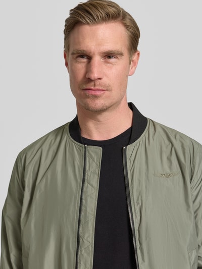 Männermodel trägt Bomberjacke mit Label-Patch und Eingrifftaschen von Original Bombers, khaki, glatte Textur, Nahaufnahme Oberkörper, Gerader-Schnitt, Reißverschluss, Reißverschlusstasche am Ärmel, Druckknopfverschluss, Gerippte-Abschlüsse, Rückenlänge bei Größe M: 67 cm, Schulterbreite bei Größe M: 43 cm, Brustweite bei Größe M: 53 cm, Ärmellänge: 68 cm.