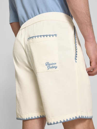 Männermodel trägt Shorts mit umstochener Kante von REVIEW, Baumwoll-Mix in Creme mit blauen Details, Bund und Tasche, Ganzkörperansicht Rückansicht.