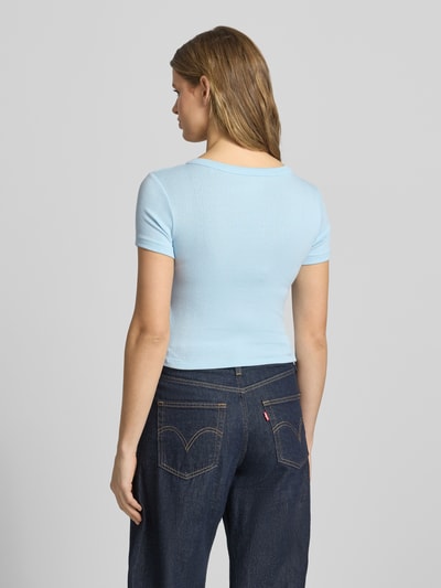 Frauenmodel trägt Cropped T-Shirt mit V-Ausschnitt und Knopfleiste von Guess Jeans in Hellblau, Material-Mix mit Viskose-Anteil, Logo, Rückenlänge bei Größe S 42 cm, Ganzkörperansicht Rückansicht.