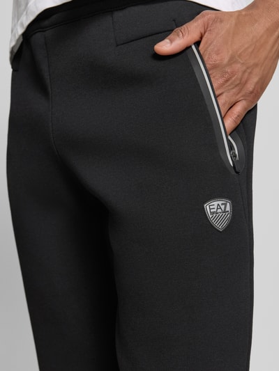 Männermodel trägt Sweatpants mit Viskose-Anteil Modell 'TRAIN PREMIUM SHIELD' von EA7 Emporio Armani, Nahaufnahme Oberschenkel. Elastischer-Material-Mix in Schwarz, Gerade Passform, Reißverschlusstaschen, gerippte Abschlüsse, Innenbeinlänge 78 cm bei Größe M.