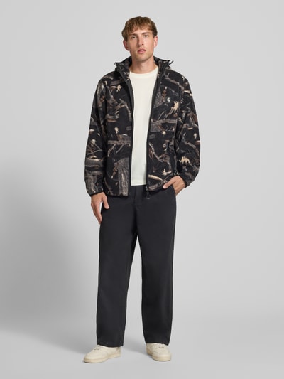 Männermodel trägt Fleecejacke mit Kapuze Modell 'Blevin' von Carhartt Work In Progress, camouflagemuster, gerader Schnitt, Zwei-Wege-Reißverschluss, Kapuze, Seitentaschen, Label-Patch, schwarze Hose, cremefarbene Schuhe, Farben in Beige, Braun, Schwarz, Ganzkörperansicht von vorne. Rückenlänge 67 cm, Ärmellänge 71 cm.